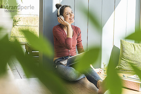 Glückliche Frau mit Kopfhörern und Laptop sitzt zu Hause am Fenster
