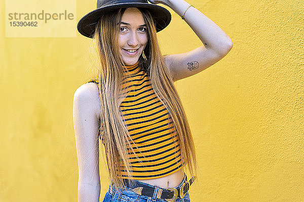 Porträt einer lächelnden Teenagerin mit Hut an gelber Wand