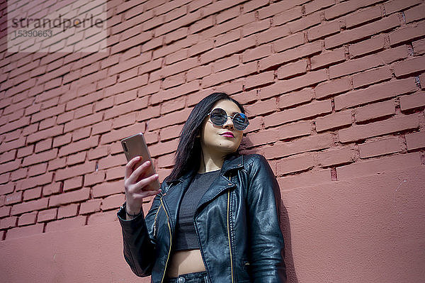 Porträt einer jungen Frau mit Smartphone  die eine Sonnenbrille und eine schwarze Lederjacke trägt