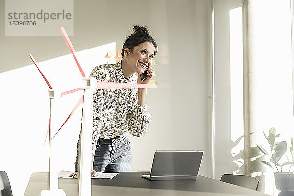 Geschäftsfrau mit Windradmodellen und Laptop auf dem Schreibtisch im Büro beim Telefonieren mit dem Handy