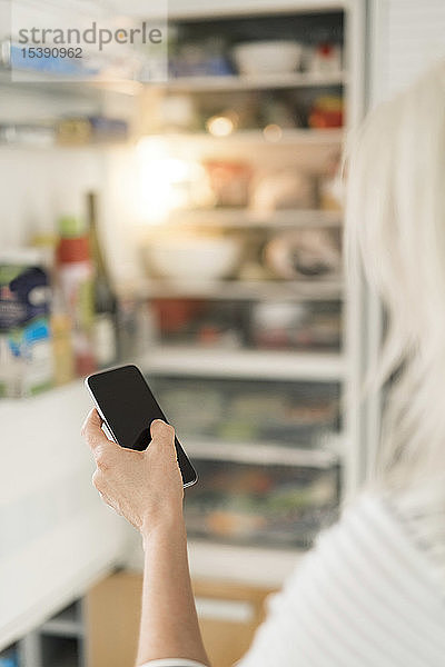 Hand einer Frau mit einem Smartphone  die den Kühlschrank in der Küche des Smart Home überprüft