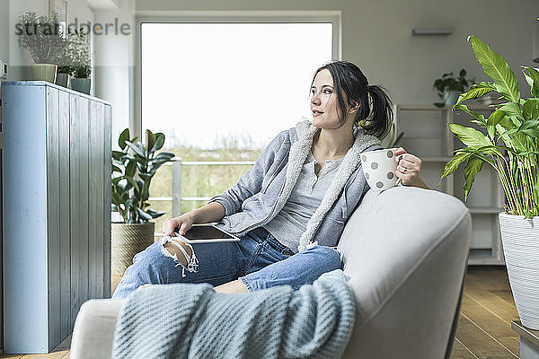 Nachdenkliche Frau mit Becher und Tablette  die zu Hause auf der Couch sitzt