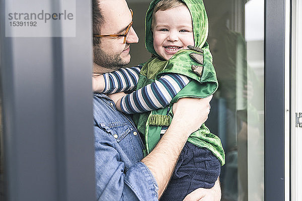 Vater trägt glücklichen Sohn in einem Kostüm an Terrassentür zu Hause