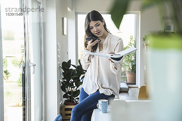 Junge Frau sitzt zu Hause am Tisch und hält Smartphone und Flugzeugmodell in der Hand