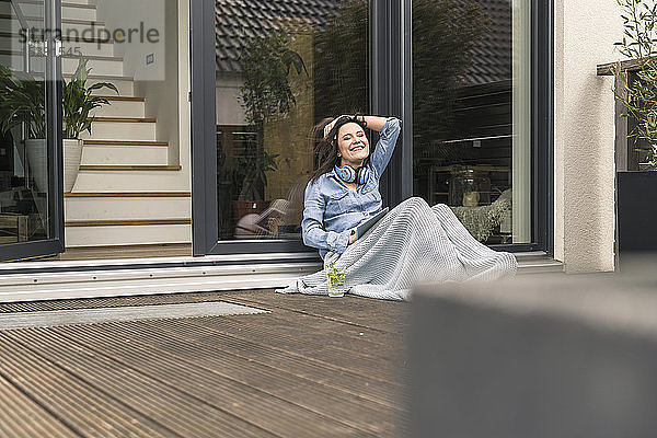 Glückliche  in eine Decke gehüllte Frau sitzt zu Hause mit Tablette auf der Terrasse