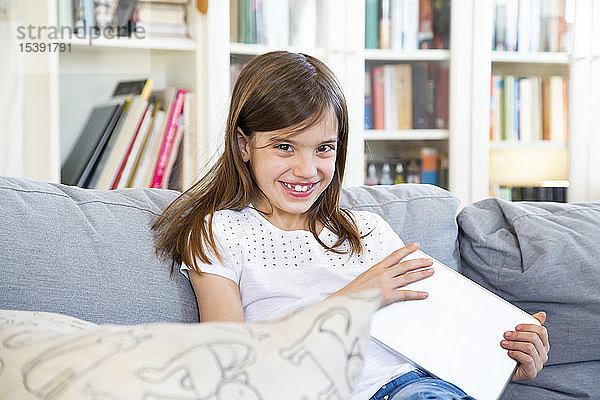 Porträt eines fröhlichen kleinen Mädchens  das zu Hause auf der Couch mit einem digitalen Tablet sitzt