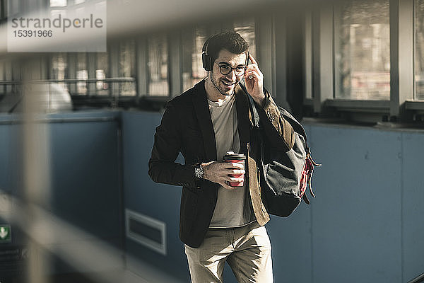 Lächelnder junger Mann mit Kopfhörern  Handy und Kaffee zum Mitnehmen auf dem Bahnhof