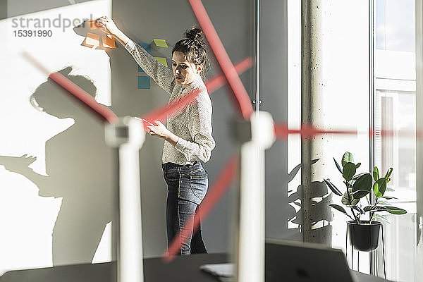 Geschäftsfrau im Büro klebt Haftnotizen an die Wand mit Modellen von Windkraftanlagen auf dem Schreibtisch
