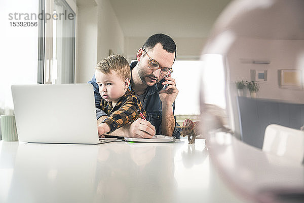 Vielbeschäftigter Vater arbeitet am Tisch im Home-Office  der Sohn sitzt auf seinem Schoß