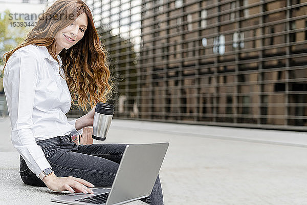 Junge Geschäftsfrau  die in der Stadt auf einer Bank sitzt  mit dem Laptop arbeitet und Kaffee trinkt