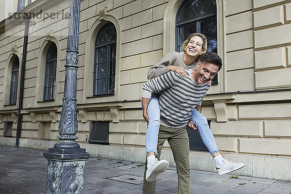 Glücklicher Mann nimmt Frau huckepack auf dem Bürgersteig in der Stadt mit