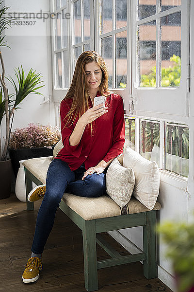 Schöne Frau sitzt auf einer Bank am Fenster und benutzt ein Smartphone