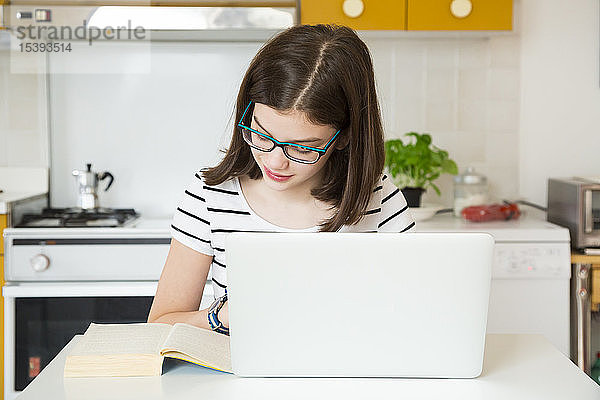 Mädchen benutzt Laptop und schaut sich in der Küche ein Buch an