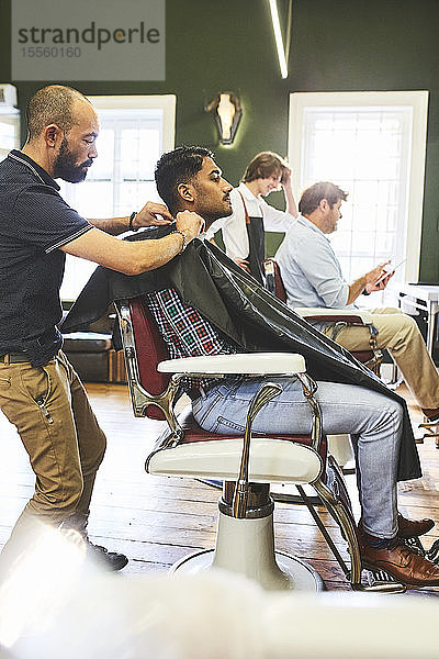 Männlicher Friseur  der einen Kunden auf einen Haarschnitt vorbereitet  in einem Friseursalon