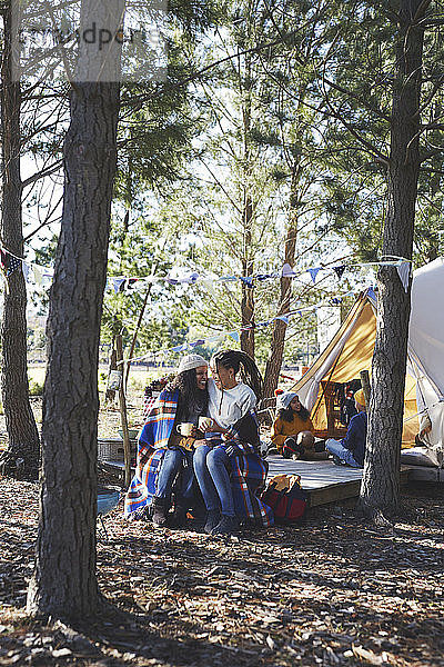 Glückliches  liebevolles lesbisches Paar mit Kindern  das sich auf einem Campingplatz im Wald entspannt und Kaffee trinkt