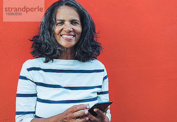 Porträt einer lächelnden  selbstbewussten Frau  die ein Smartphone vor rotem Hintergrund benutzt
