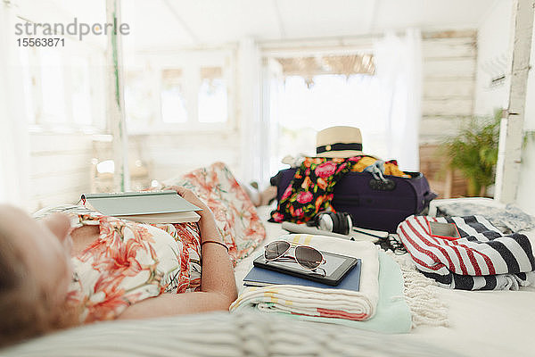 Frau mit Buch entspannt auf Bett neben Koffer in sonnigen Strandhütte Schlafzimmer