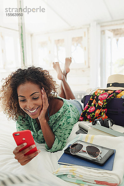 Junge Frau mit Smartphone entspannt auf dem Bett neben Koffer und Habseligkeiten