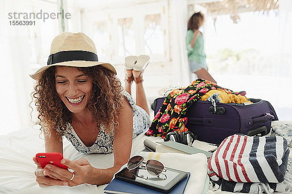 Glückliche junge Frau benutzt ihr Smartphone und packt ihren Koffer auf dem Bett der Strandhütte aus