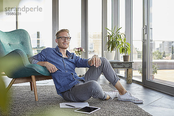 Lächelnder junger Mann sitzt auf Teppich mit Tablette und Notizblöcken zu Hause