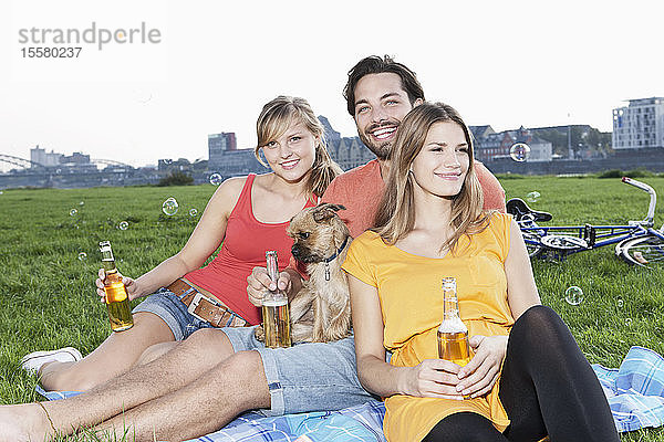Deutschland  Köln  Junger Mann und Frau mit Hund und Bierflasche im Gras  lächelnd