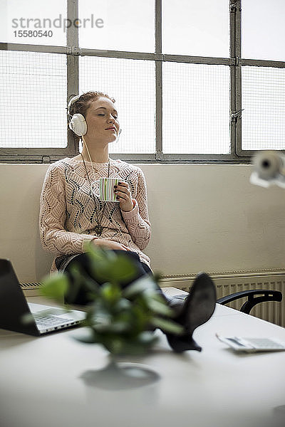 Junge Frau sitzt am Schreibtisch mit Kopfhörern und einer Tasse Kaffee