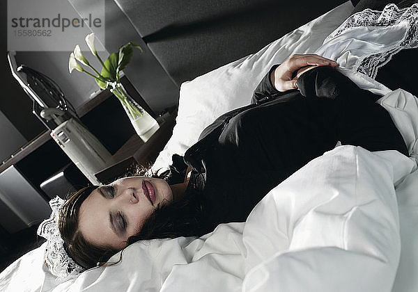 Deutschland  Junge Frau als Dienstmädchen verkleidet auf dem Bett liegend