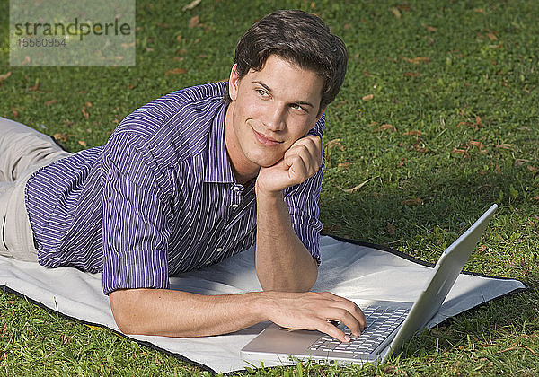 Deutschland  München  Junger Mann im Gras liegend und mit Laptop