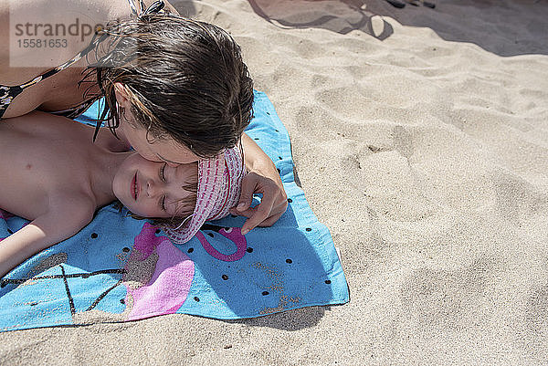 Mutter küsst Tochter  die auf einem Strandtuch am Strand liegt