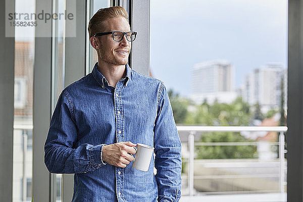 Lächelnder junger Mann mit einer Tasse Kaffee an der Balkontür stehend