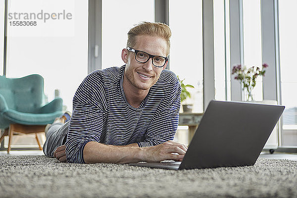 Porträt eines lächelnden jungen Mannes  der zu Hause mit seinem Laptop auf dem Teppich liegt