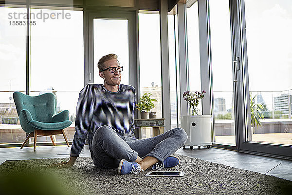 Lächelnder junger Mann sitzt zu Hause auf Teppich mit Tablette