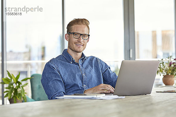 Porträt eines lächelnden jungen Mannes  der mit einem Laptop am Tisch sitzt