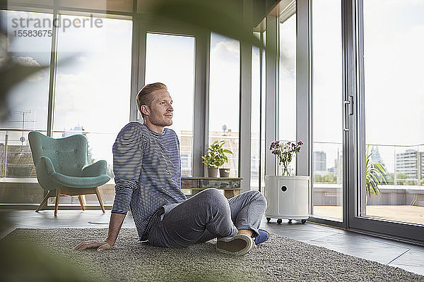 Junger Mann sitzt zu Hause auf Teppich und schaut aus dem Fenster