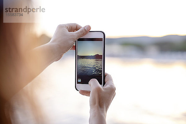 Nahaufnahme einer Frau  die ein Handyfoto vom Meer bei Sonnenuntergang macht