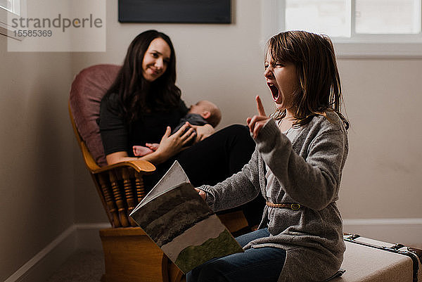 Mädchen mit Buch  das Gesichter zieht  während die Mutter den kleinen Bruder im Wohnzimmersessel wiegt
