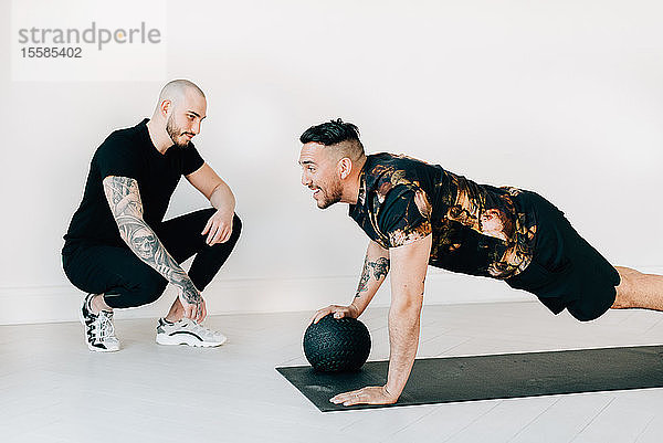 Fitnesstrainerin beobachtet Mann  der im Studio ein Brett mit Medizinball bearbeitet