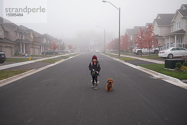 Mädchen geht mit dem Hund inmitten einer nebligen Vorstadtstraße spazieren  Portrait in voller Länge