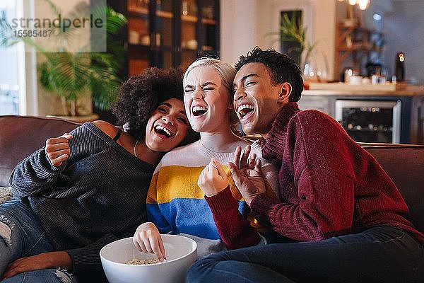 Junger Mann und junge Freundinnen lachen beim Fernsehen auf dem Wohnzimmersofa mit einer Schüssel Popcorn