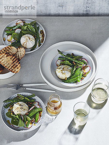 Stilleben in hoher Tonart mit Weißweingläsern und Tellern mit Burrata-Frühlingssalat auf weißem Tisch  Draufsicht