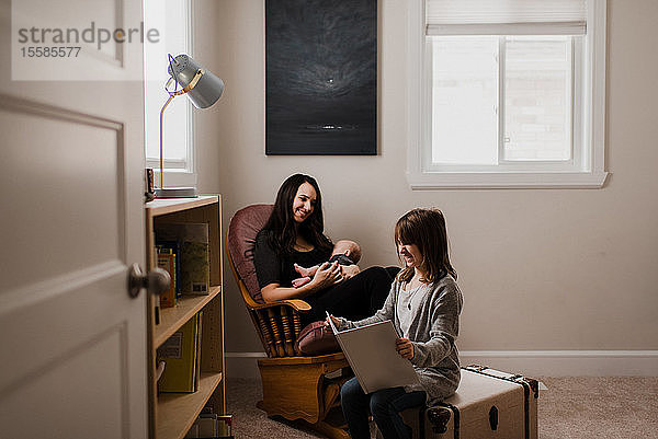 Junge Frau hält den kleinen Sohn im Wohnzimmersessel  während die Tochter ein Buch liest