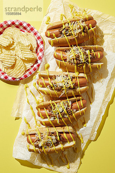 Reihe von Coney-Hotdogs mit geträufeltem Senf und einem Teller Kartoffelchips auf gelbem Hintergrund  Draufsicht
