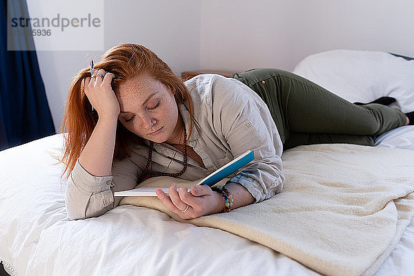 Junge Frau mit roten Haaren liegt auf dem Bett und liest ihr Tagebuch