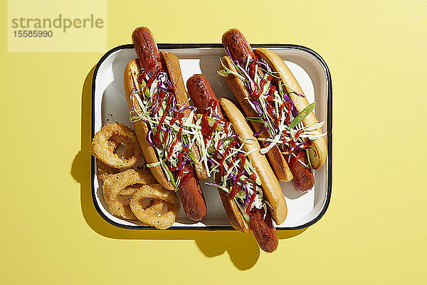 Drei Hotdogs auf Tablett mit Zwiebelringen  Draufsicht auf gelben Hintergrund