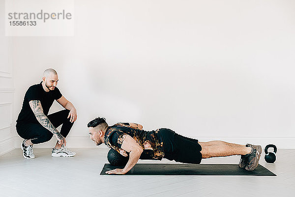 Fitnesstrainerin beobachtet Mann  der im Studio ein Brett mit Medizinball bearbeitet