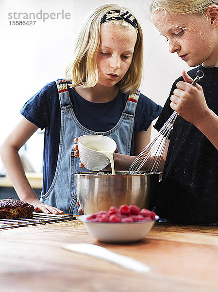 Mädchen und ihre Schwester backen einen Kuchen und gießen Sahne in eine Rührschüssel am Küchentisch