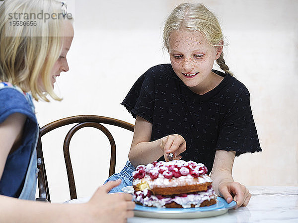 Mädchen und ihre Schwester schneiden ihren selbstgebackenen Kuchen mit frischen Himbeeren am Küchentisch an