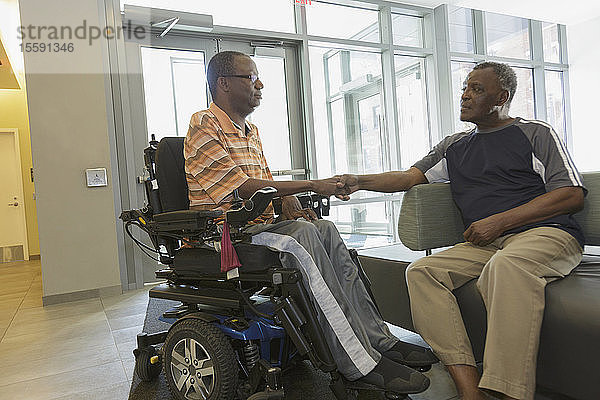 Mann mit Guillain-Barre-Syndrom auf einem elektrischen Stuhl gibt seinem Vater im Wohnzimmer die Hand