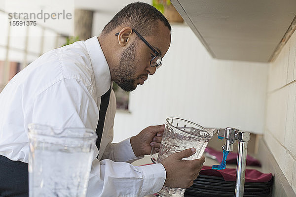 Afroamerikanischer Mann mit Down-Syndrom als Kellner  der in der Küche einen Krug mit Wasser auffüllt