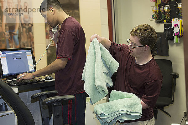 Junger Mann mit Down-Syndrom bei der Vorbereitung von Handtüchern in der Ausrüstungsabteilung einer Hochschule für das Fitnessstudio mit einem Betreuer am Telefon